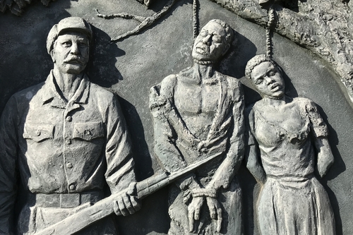 Steinstatuen in Wand gehauen. Weißer Mann mit Gewehr, daneben ein gehängtes afrikanisches Ehepaar