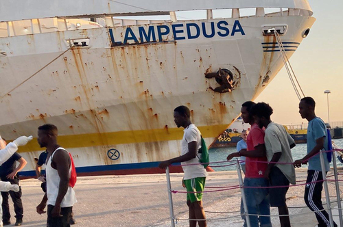 Bug eines Schiffes mit Namen Lampedusa im Hafen. Im Vordergrund Geflüchtete