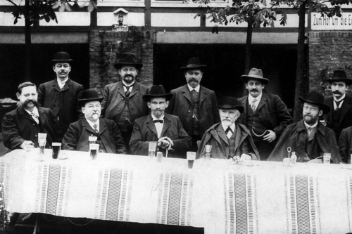 Schwarz-Weiß-Fotografie von elf Männern in Anzügen mit Hut, für ein Gruppenfoto an einem Tisch vor einem Ausflugslokal (Fachwerkhaus) posierend