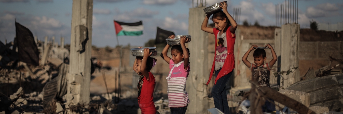 Drei Mädchen laufen über die zerbombten Trümmer von Häusern und tragen Metallschüsseln auf ihrem Kopf, die sie mit beiden Händen festhalten. Im Hintergrund weht eine palästinensische Flagge.