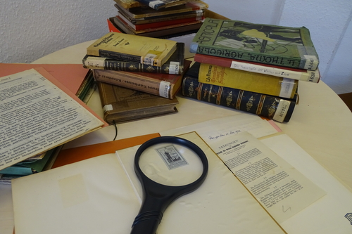 Stapel mit Bibliotheksbüchern auf einem Tisch, im Vordergrund ein aufgeschlagenes Buch, auf dem eine Lupe liegt, die auf dem auf der Innenseite angebrachten Buch-Siegel liegt.