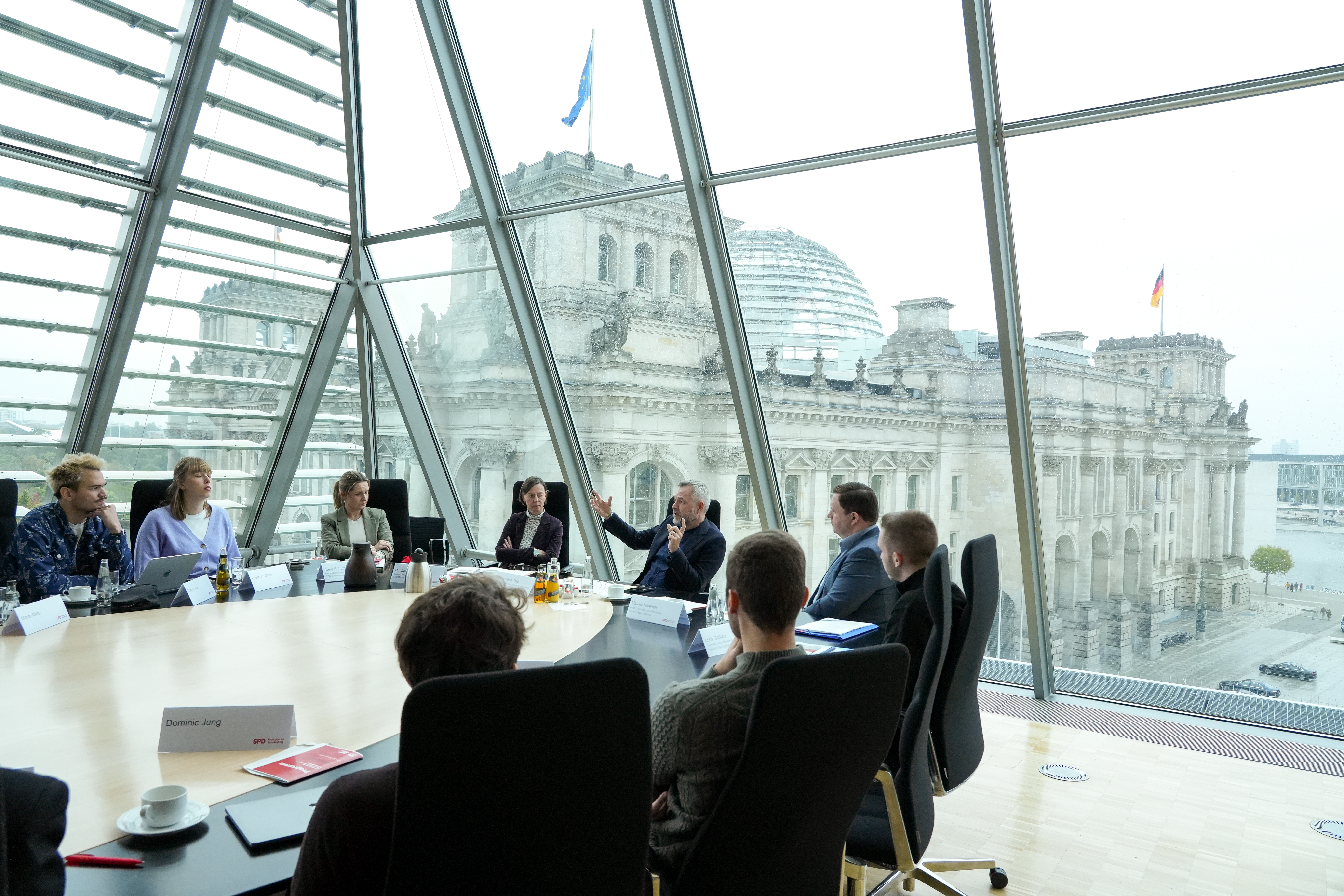 Teilnehmer_innen diskutieren mit Dr. Michael Roth an einem großen Tisch. Im Hintergrund ist der Bundestag zu sehen.
