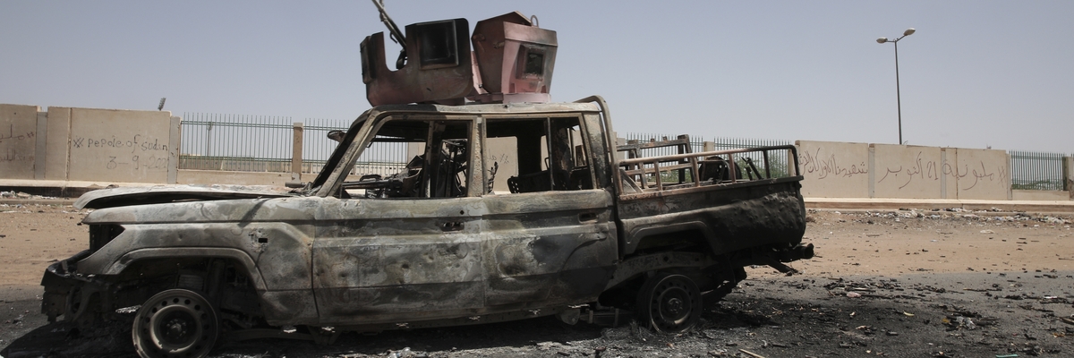 Ein zerstörtes Militärfahrzeug, vermutlich ausgebrannt, steht vor einem Gelände mit hohem Zaun, der an den Seiten in Betonmauern übergeht. Die Szene ist menschenleer.
