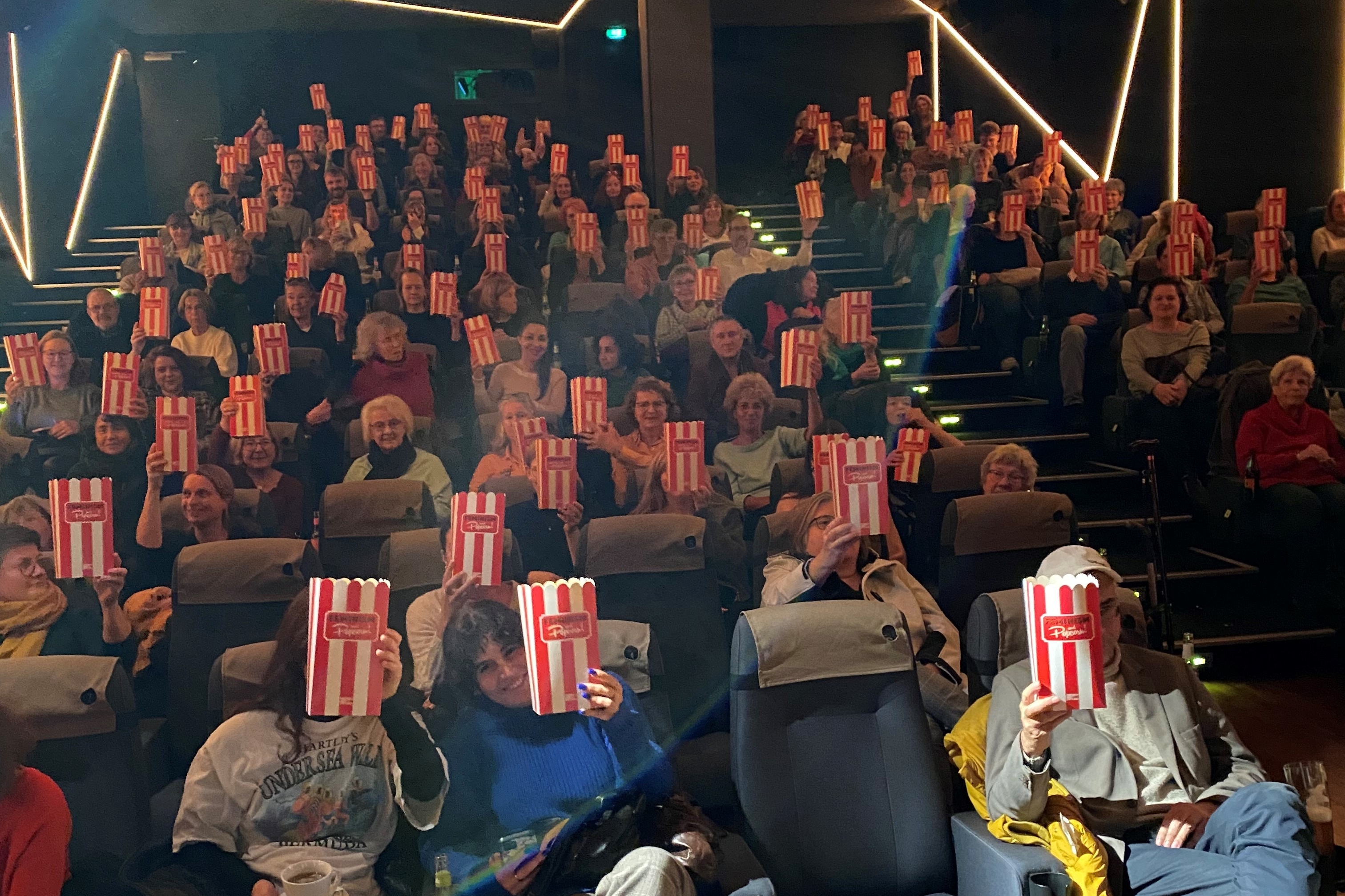 gefüllter Kinosaal, alle halten eine Popcorntüte in die Luft