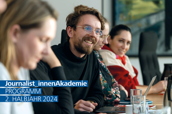 Eine Gruppe Seminarteilnehmer_innen sitzt an einem Tisch. Im Fokus ein junger Mann, der aufmerksam lächelt.