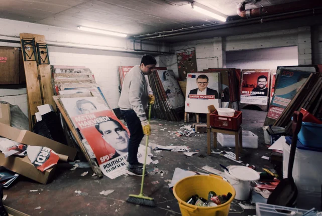 Person steht zwischen alten Wahlplakaten in einem Keller mit einem Besen und fegt