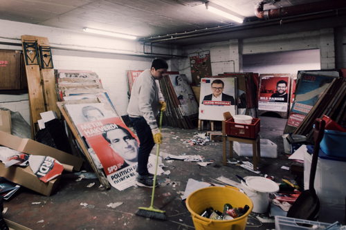 Person steht zwischen alten Wahlplakaten in einem Keller mit einem Besen und fegt