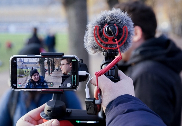 Eine Person filmt mit einem Smartphone mit aufgestecktem Mikrophon zwei sich unterhaltende Personen.