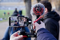 Eine Person filmt mit einem Smartphone mit aufgestecktem Mikrophon zwei sich unterhaltende Personen.
