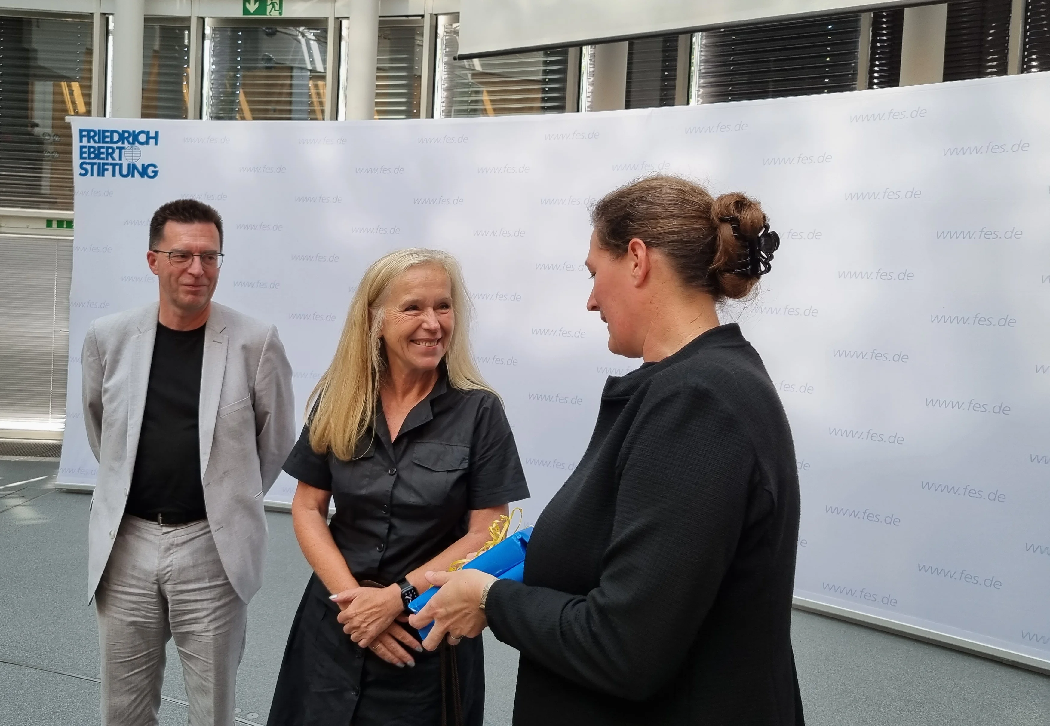 Geschenkübergabe von Anne Haller an Susanne Lang mit Serge Embacher im Hintergrund