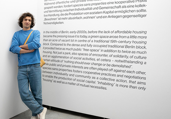 Eine Person steht mit dem Rücken die Wand gelehnt. Auf der Wand sind Ausstellungstexte zu sehen.