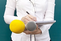 Frau mit weißer Bluse hält zwei Mikrophone und schreibt etwas auf einen Notizblock.