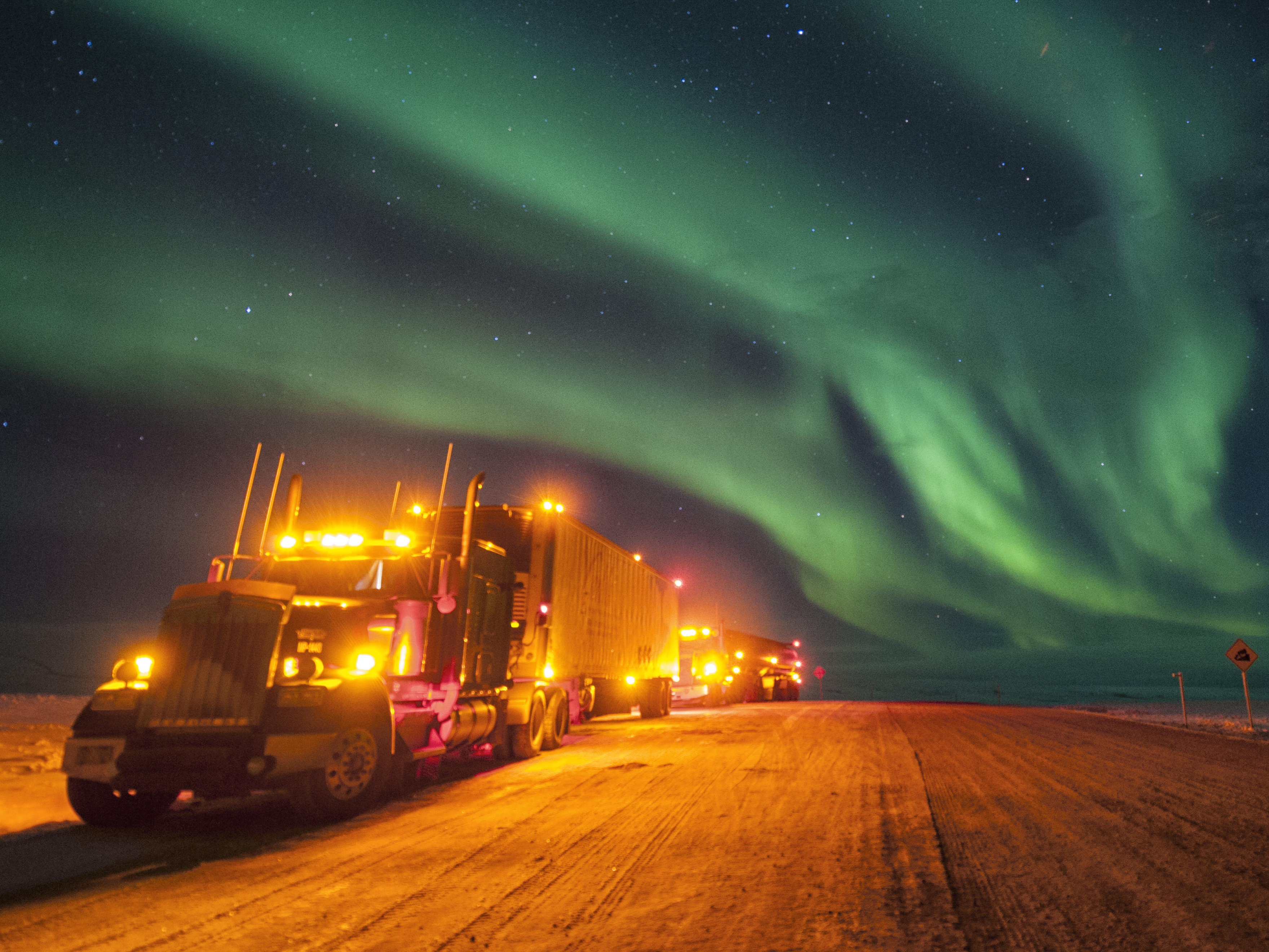 Beleuchteter LKW in einer winterlichen Landschaft im Dunkeln. Am Himmel sind Nordlichter zu sehen.