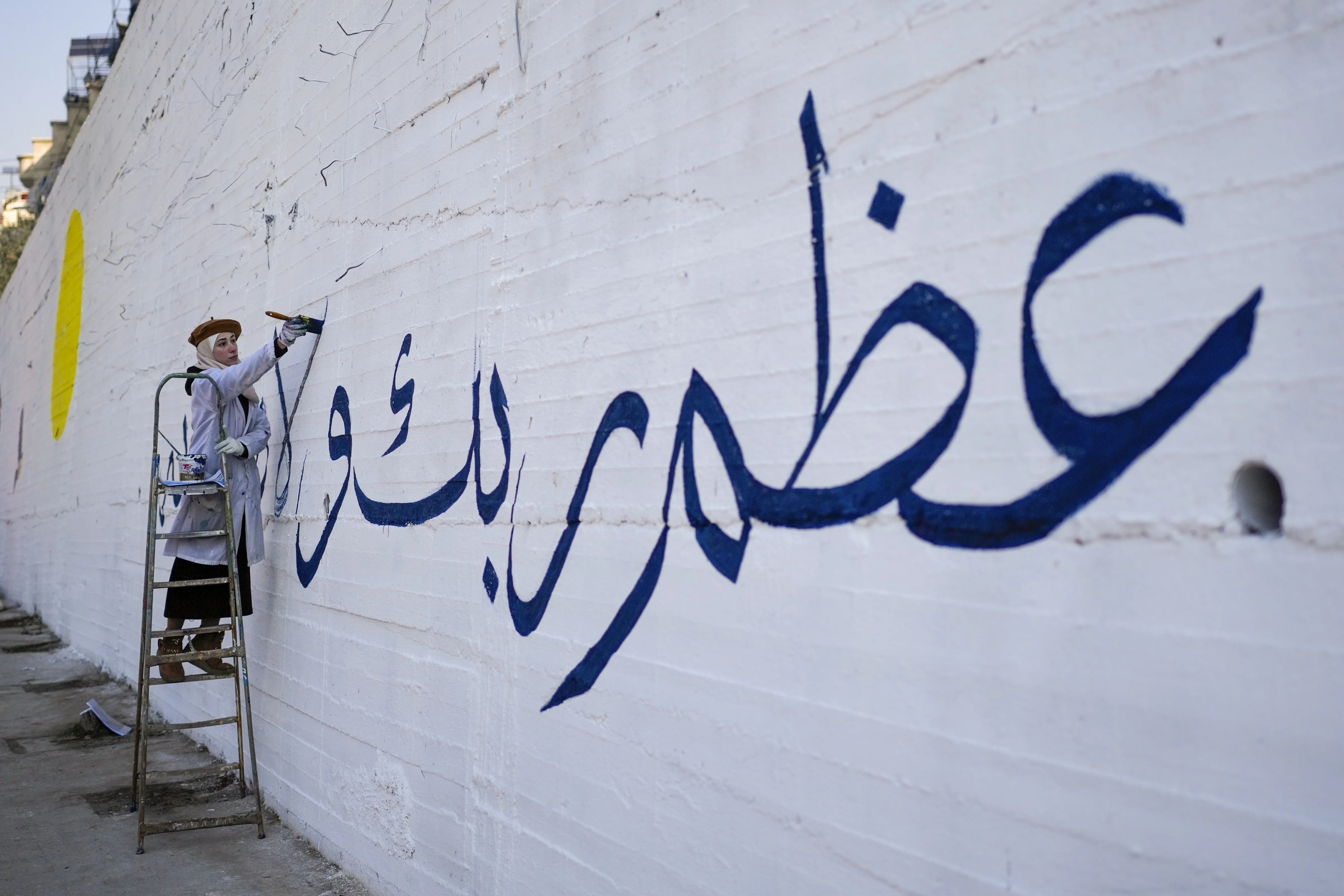 Eine Frau schreibt "Betet euren Gott an" auf Arabisch, während eine Gruppe junger Freiwilliger ein Wandbild mit dem Symbol des Friedens an eine Mauer am Stadtrand von Damaskus malt.