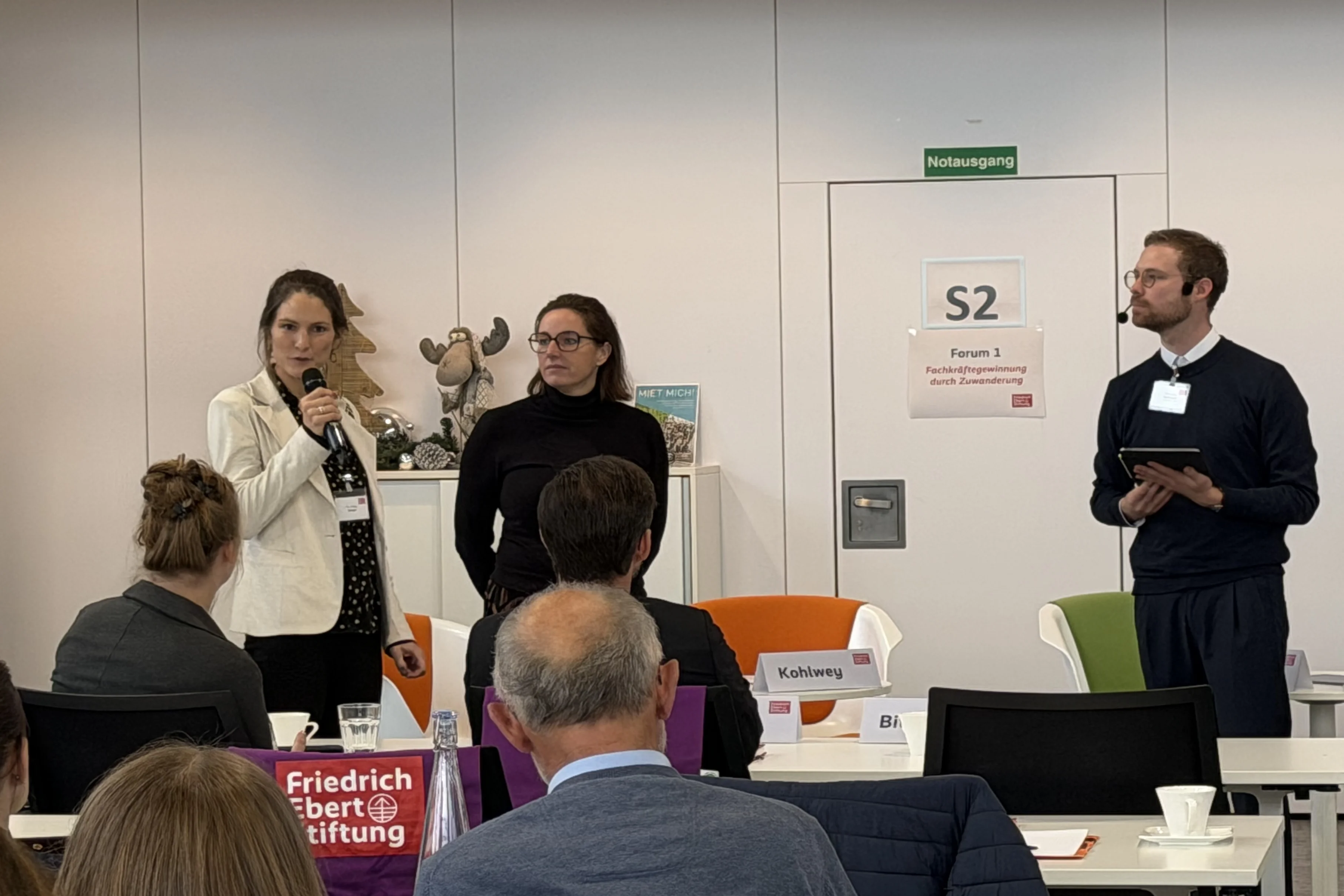 Mehrere Personen stehen und sitzen in einem Konferenzraum, eine Person spricht mit Mikrofon, im Vordergrund sind Namensschilder und ein Schild mit 'Friedrich Ebert Stiftung' sichtbar.