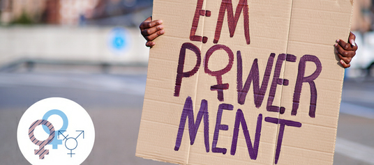 Person hält ein handgemachtes Plakat mit der Aufschrift 'EMPOWERMENT' in lila und roter Farbe, wobei das 'M' und 'W' besonders hervorgehoben sind. Der Hintergrund ist unscharf mit städtischer Kulisse.