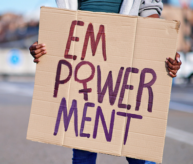 Person hält ein handgemachtes Plakat mit der Aufschrift 'EMPOWERMENT' in lila und roter Farbe, wobei das 'M' und 'W' besonders hervorgehoben sind. Der Hintergrund ist unscharf mit städtischer Kulisse.