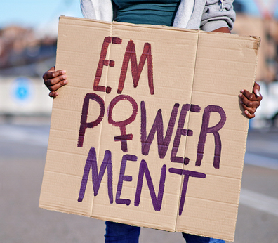 Person hält ein handgemachtes Plakat mit der Aufschrift 'EMPOWERMENT' in lila und roter Farbe, wobei das 'M' und 'W' besonders hervorgehoben sind. Der Hintergrund ist unscharf mit städtischer Kulisse.