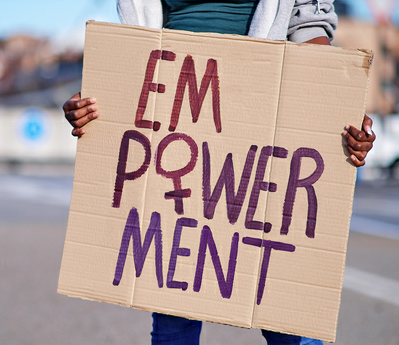 Person hält ein handgemachtes Plakat mit der Aufschrift 'EMPOWERMENT' in lila und roter Farbe, wobei das 'M' und 'W' besonders hervorgehoben sind. Der Hintergrund ist unscharf mit städtischer Kulisse.