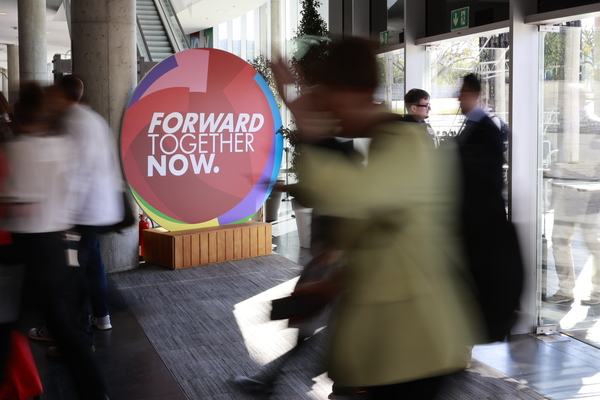 verschommen aufgenommene Menschen gehen vor einem runden SChild vorbei mit der Aufschrift: Foward Together. Now.