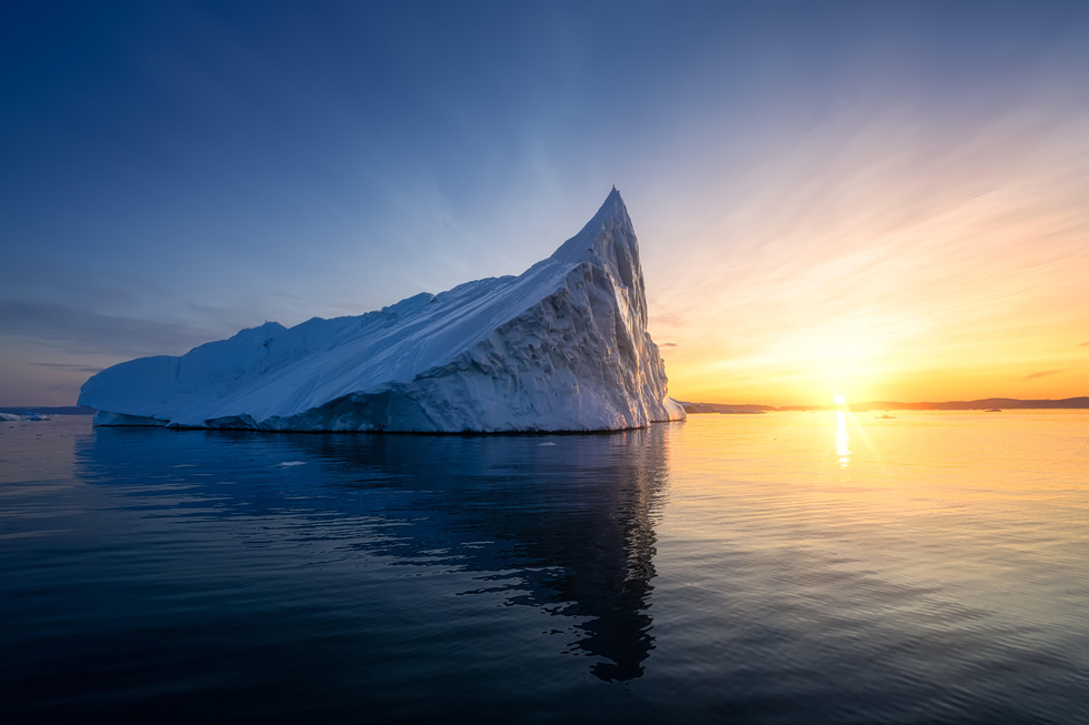 Ein Eisberg im Meer, im Hintergrund geht die Sonne unter.