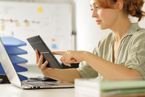 Eine junge Frau arbeitet konzentriert in einem Büro am Schreibtisch und betrachtet ein Tablet und einen Laptop.