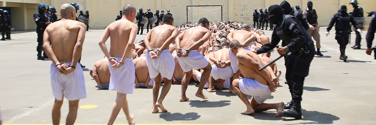 100 männliche Häftlinge in El Salvador sitzen eng beinander auf einm kahlen Boden und sind mit Handschellen gefesselt.