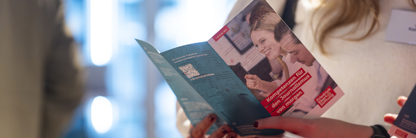 Der Programmflyer der Journalist:innen-Akademie der FES mit der Aufschrift "Kompetenzen für den Journalismus von morgen" wird von einer jungen Frau gelesen.