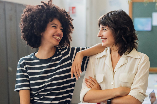 Zwei Frauen stehen nebeneinander und lächeln sich an, vor einem Büro-Hintergrund