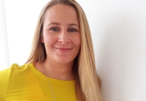 Porträt der Trainer_in Vivian Upmann, die in einem gelben Oberteilt vor einer weißen Wand steht.