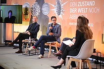 Ein Mann sitzt mit anderen Menschen auf einem Podium und hält ein Mikrofon in der Hand.