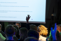 Foto vom Publikum bei einer Podiumsdiskussion; einige Personen heben die Hand, im Hintergrund ist eine Leinwand mit Programminformationen zu sehen.