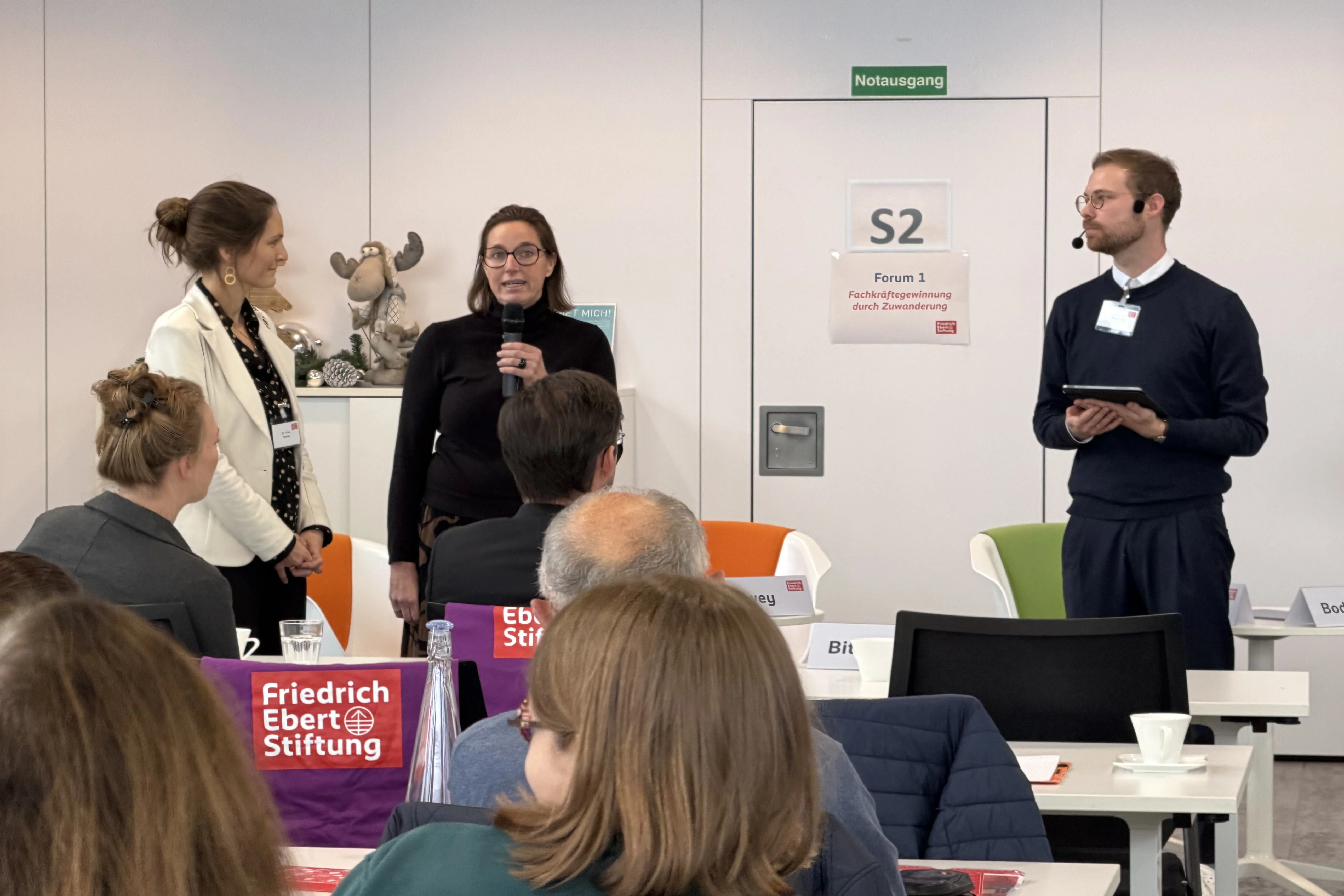 Mehrere Personen stehen und sitzen in einem Konferenzraum. Drei Frauen stehen vorne, eine hält ein Mikrofon. Ein Mann steht rechts mit einem Tablet. Auf Tischen liegen Schilder mit 'Friedrich Ebert Stiftung'.