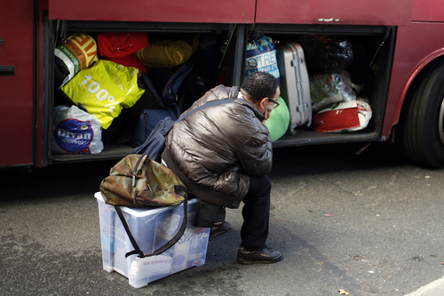 Ein Mensch sitzt vor einem Bus auf einer Kiste mit seinen Habseligkeiten. Das Gepäckfach des Buses steht offen und ist voll mit Gepäck.