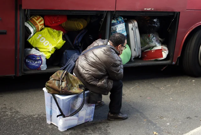 Ein Mensch sitzt vor einem Bus auf einer Kiste mit seinen Habseligkeiten. Das Gepäckfach des Buses steht offen und ist voll mit Gepäck.