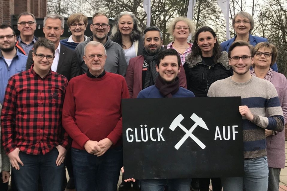 Viele Personen stehen nebeneinander und hintereinander. In der vorderen Reihe halten zwei Männer ein schwarzes Schild hoch, auf dem steht: "Glück auf".