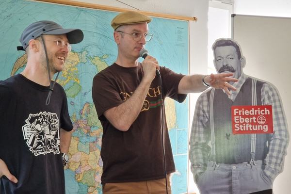 Brian Vit, Tony L. und eine Friedrich-Ebert-Pappfigur stehen vor einer Weltkarte, Tony spricht in ein Mikro zu einem unsichtbaren Publikum