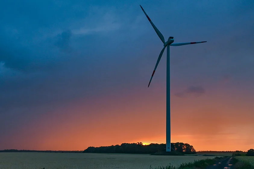 Ein Windrad an einem Feldweg, im Hintergrund ein Sonnenuntergang hinter einer Gewitterwolke