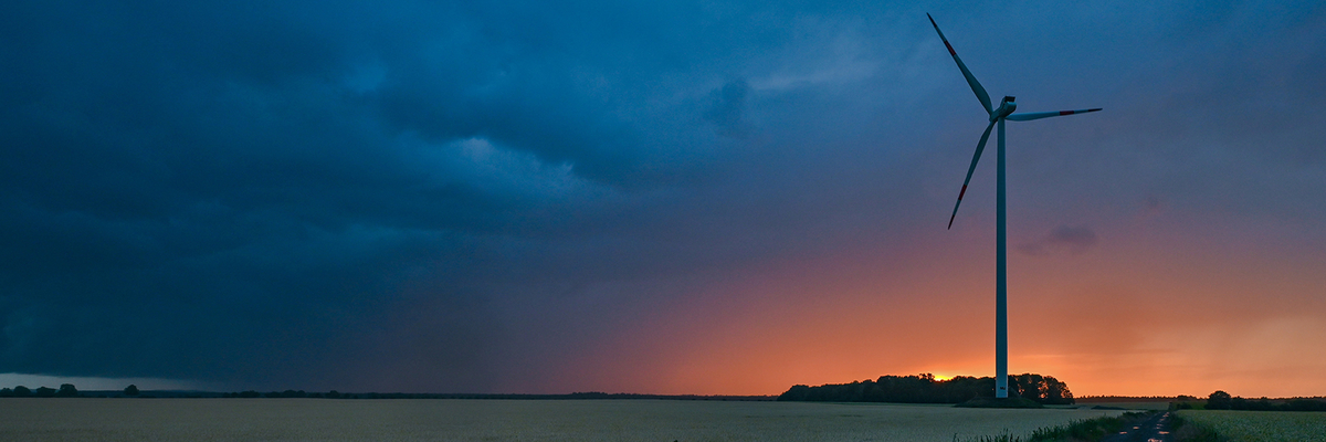 Ein Windrad an einem Feldweg, im Hintergrund ein Sonnenuntergang hinter einer Gewitterwolke