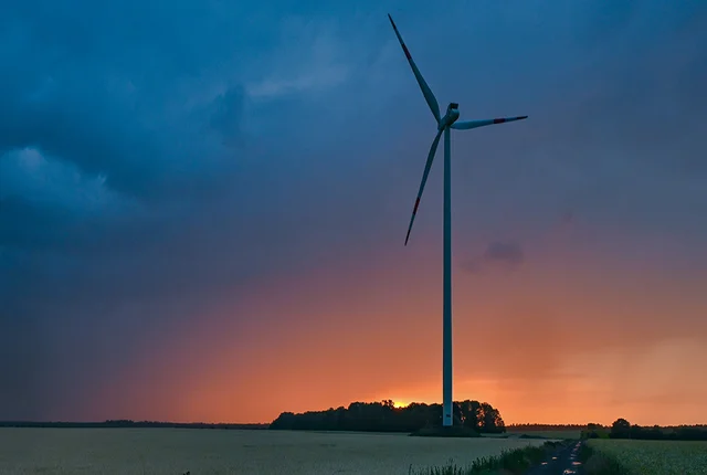 Ein Windrad an einem Feldweg, im Hintergrund ein Sonnenuntergang hinter einer Gewitterwolke