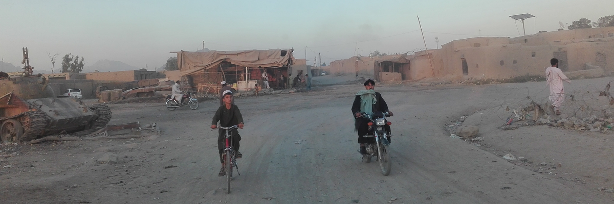 Ein Kind auf einem Fahrrad und ein Mann auf einem Moped in einem afghanischen Dorf bei Kandahar, links ist ein Panzer zu sehen. 