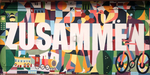 Schriftzug "Zusammen" in Großbuchstaben auf einem buntbemalten Tor mit Motiven des Zusammenlebens wie menschlichen Figuren, einem Fahrrad, Bäumen