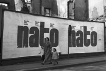 Frau mit Kind gehen über die Straße. Vor einem zerstörten Gebäude hängt ein Plakat, das an das 10-jährige Jubiläum der NATO erinnert.