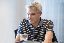 Portrait von Trainer Bernd Oswald, der in einem blau-weiß gestreiften Poloshirt an einem Tisch im Seminarraum sitzt.