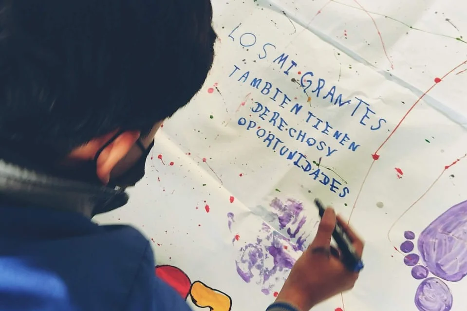 Ein Kind schreibt und malt auf ein Transparent. Auf Spanisch ist zu lesen "Auch Migrant:innen haben Rechte und Chancen."