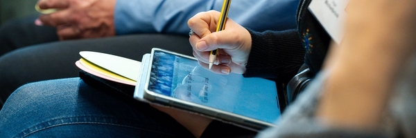 Foto einer sitztenden Person, die mit einem Eingabestift auf ein Tablet schreibt.