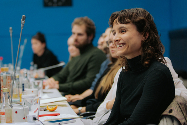 Foto: Mehrere junge Leute sitzen in einer Reihe an einem Tisch, der Fokus liegt auf einer lächelnden Frau.