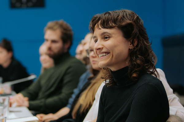 Foto: Mehrere junge Leute sitzen in einer Reihe an einem Tisch, der Fokus liegt auf einer lächelnden Frau.