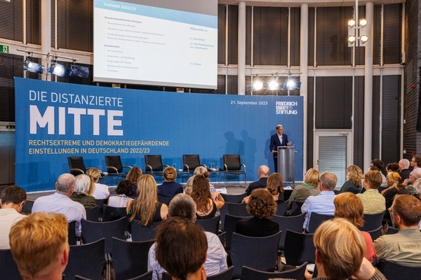 Ein Mann steht auf einer Bühne und spricht vor Publikum. Auf der Podiumsrückwand ist „Die distanzierte Mitte“ zu lesen.