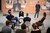 Teilnehmer_innen der MedienAkademie sitzen in einem Stuhlkreis und eine Teilnehmerin spricht in ein Mikrophon.